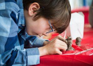 A close up of a young boy with brown hair from the side wearing a blue flannel shirt and glasses. He is leaning over and working intently on a clay creation that is on the table in front of him. The table has a bright red table cloth on it.