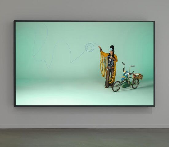 A photograph of a lightbox artwork by Dana Claxton. The lightbox has a black frame and is on a white wall. The artwork photograph is of a man in Indigenous clothing, standing next to a bike and throwing a lasso. He stands in front of an indistinct aquamarine backdrop.