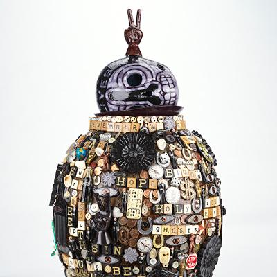 A photograph of an artwork by Jan Wade. It is a vase with lid covered in scrabble tiles and other ephemera.