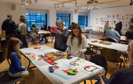 A room of people doing making activities. In the foreground, there is a white table with art supplies on it, and 2 girls with a woman working on a project.