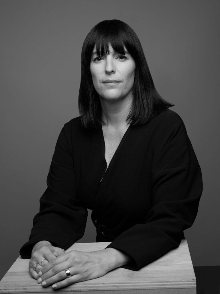 Portrait of Eva Respini in black hair. The photograph is of a woman with brown, shoulder length hair and bangs, from the torso up. Her hands are folded in front of her on a box, and she is wearing a black suit.