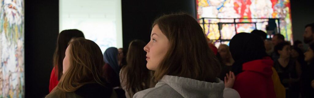 A close-up of a teenage girl's profile as she stands in a room of art and other teens, enjoying artworks by Takashi Murakami