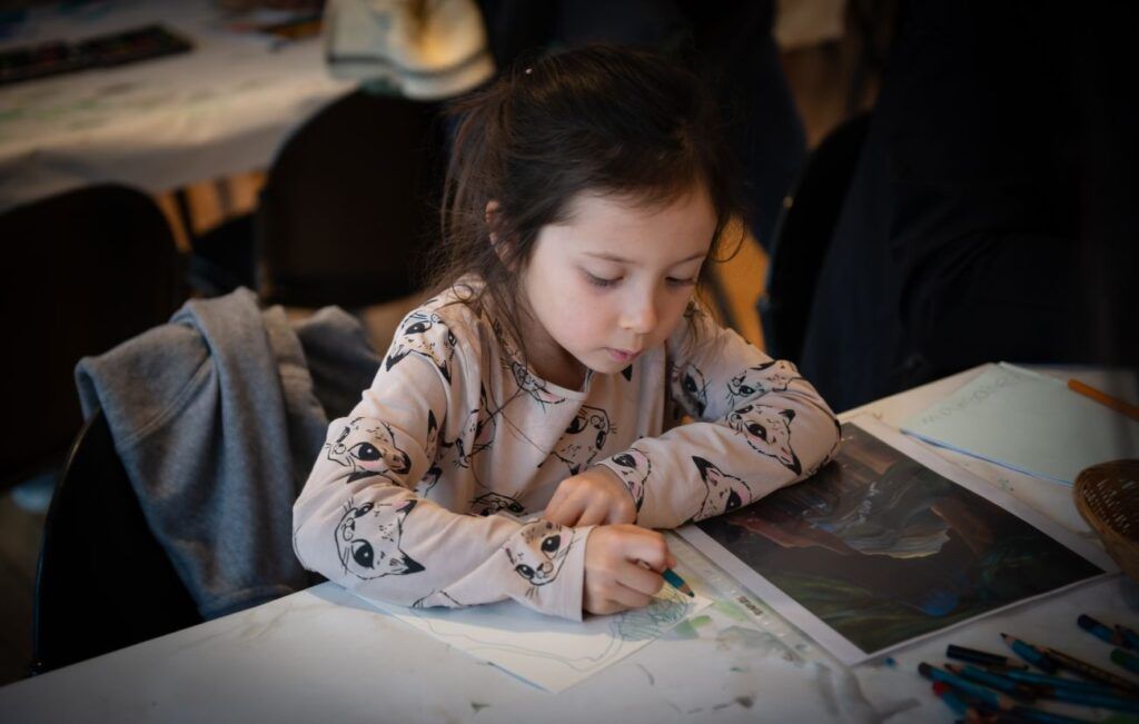 A photograph of a young girl sitting at a table and drawing.