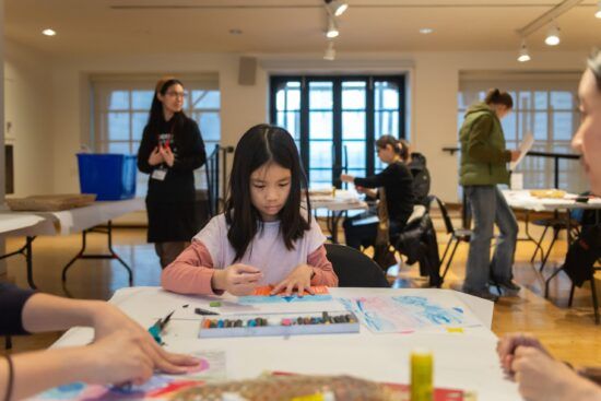 A photograph of a young girl at the Gallery in the Making Place studio. She has pastels in front of her and is drawing. She sits at a table covered in white paper.