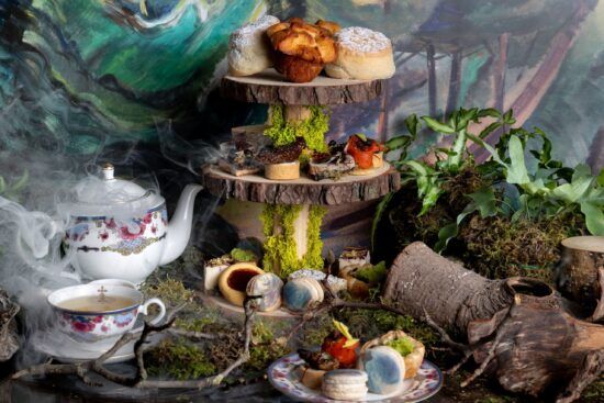 A photograph of an enchanted high tea inspired by the paintings of Emily Carr. There is a tea pot and cups to one side and at the centre is a cornucopia of tiny delights all placed on a three-tiered tray made of cut logs.