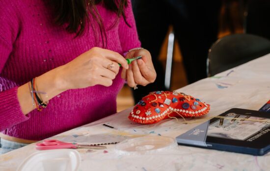 Detail of a woman's hands as she strings beads on a thread to sew onto a felt heart, sitting on the table in front of her.