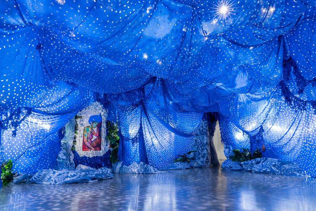 An room decorated with fabrics, faux plants and various materials and the ceiling is draped with blue fabrics with holes cut into them to resemble the night sky. A painting of a woman hangs on the wall.