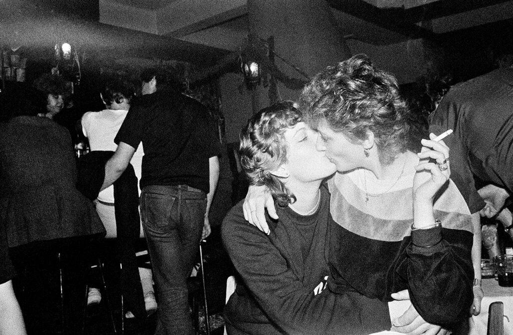 A black and white photograph of two individuals kissing int he foreground, standing in a dark nightclub.