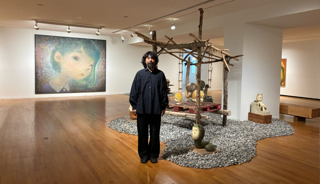Otani Workshop stands in a room of his exhibition "Otani Workshop: Monsters in My Head" at the Vancouver Art Gallery. A painting of a girl and an installation of rocks and a pagoda-like structure made of sticks are visible behind him.