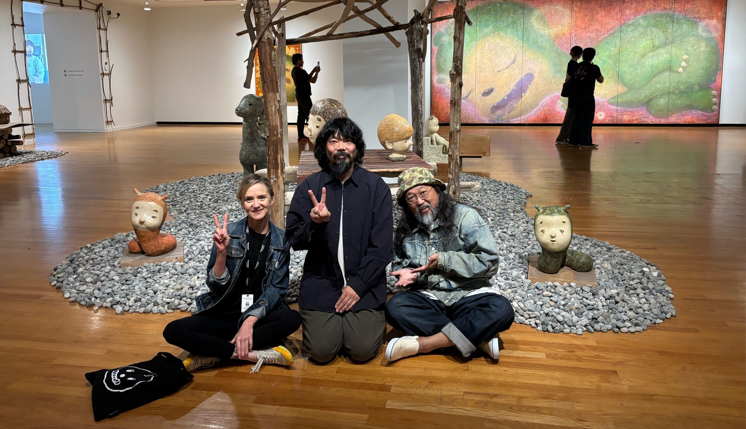 Diana Freundl, Senior Curator, Otani Workshop and Takashi Murakami sit in a room of the exhibition "Otani Workshop: Monsters in My Head" at the Vancouver Art Gallery. A painting of a sleeping dinosaur and an installation of rocks and a pagoda-like structure made of sticks are visible behind them.