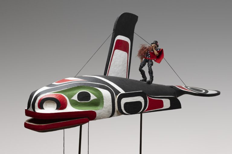 Photograph of an Indigenous installation artwork by Beau Dick of a large wooden whale with a small doll-like figurine of the artist on the whale's back.