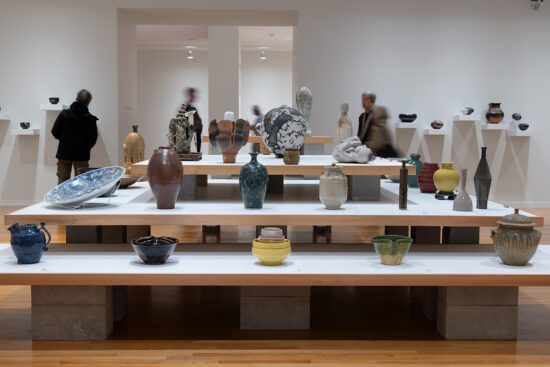 An installation view of Written In Clay. It looks at 3 shelves from the side. The shelves have multiple colourful vases and ceramics on them.