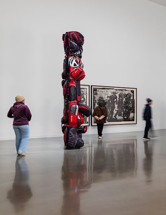 Installation view of the exhibition Postcards from the Heart: Selections from the Brigitte and Henning Freybe Collection. A sculpture that looks like a totem pole is visible in the foreground. It is made out of red golf bags. A woman stops to look at it, and two other visitors are visible in the background.