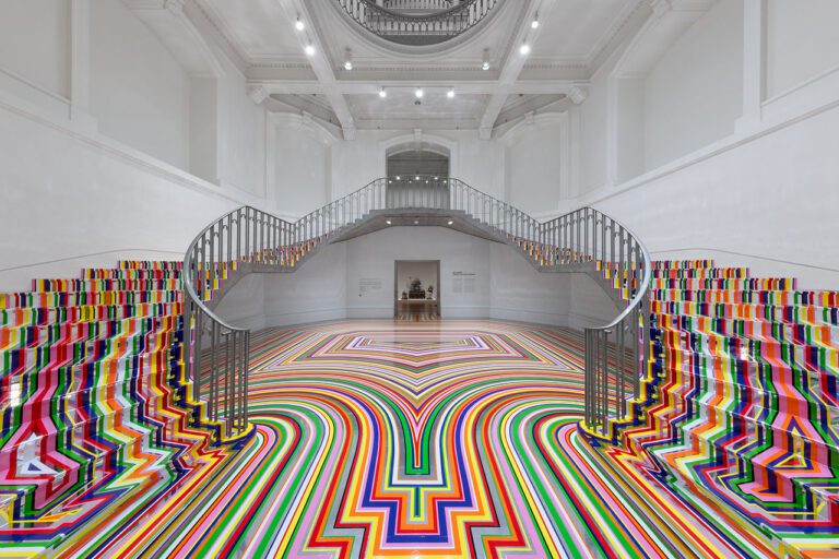 An installation photograph of Jim Lambie's artworks Zobop (Colour-Chrome) and Zobop (Colour-Chrome) Stairs covering the stairs and floor of the Vancouver Art Gallery's neo-classical Rotunda. The floor and stairs are covered in brightly coloured strips of vinyl in purple, pink, green, red, orange and yellow.