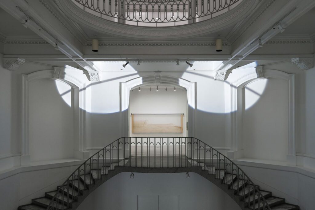 Installation view of Lucy Raven: Murderers Bar, exhibition at the Vancouver Art Gallery, April 13 to September 28, 2025. The image shows the Gallery's Rotunda, with two spotlights closing in on a opening, where a second artwork is visible on the landing.