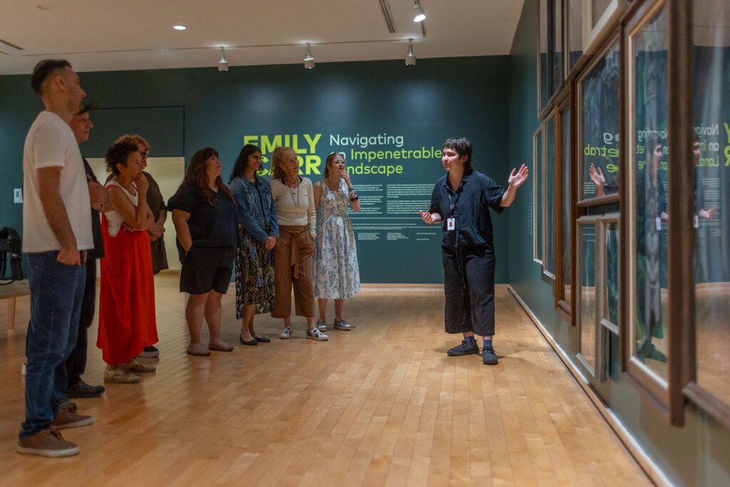 A group of men and women visit the Gallery and enjoy a guided tour. In this photograph, we can see them standing from the side, looking at a dark green wall with Emily Carr paintings of the forest on it. A young woman tour guide stands in front of the paintings gesturing and looks at the group.