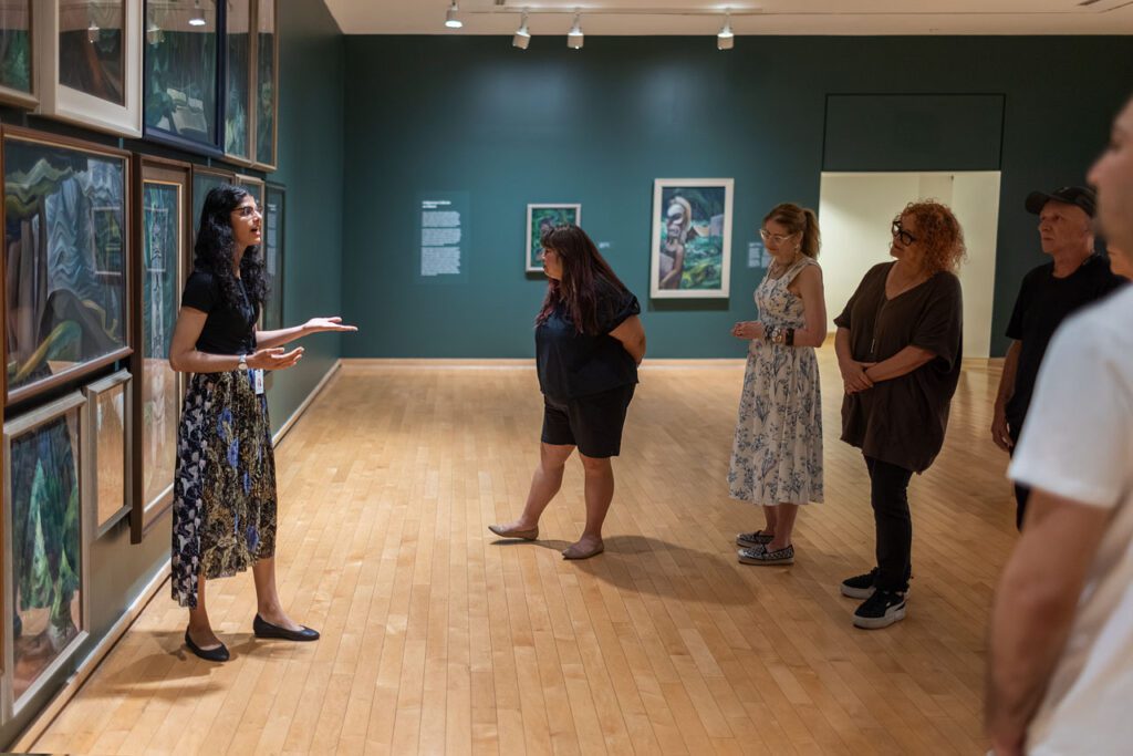A group of women and men visit the Gallery and enjoy a guided tour. They are standing looking at a dark green wall with Emily Carr paintings of the forest on it. A young woman tour guide stands in front of the paintings gesturing and speaking to the group.