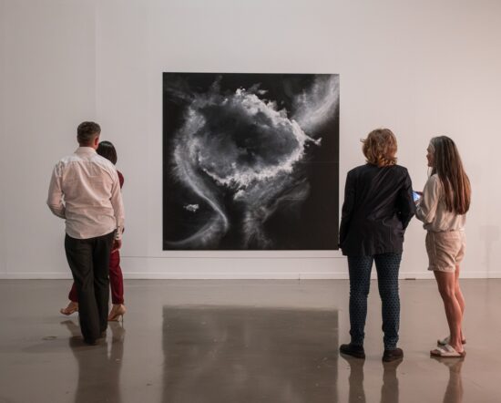 A photograph of 4 people (two women standing together and a man and a woman) from behind looking at Tacita Dean's pastel drawing of clouds in shades of black and white and grey.