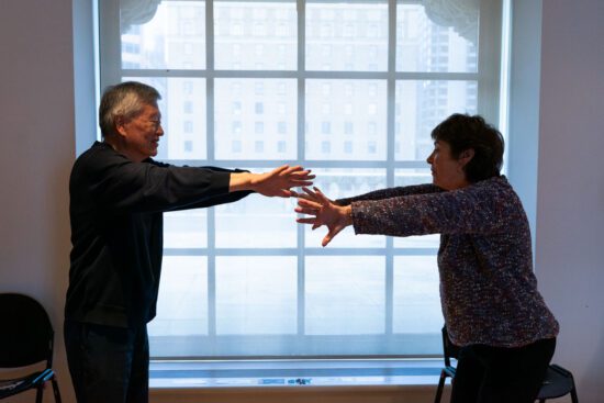 A photograph of two seniors from the side, one man on the left and one woman on the right. They are standing in front of a window grid with their arms stretched out towards each other, hands almost touching, as part of a movement workshop.