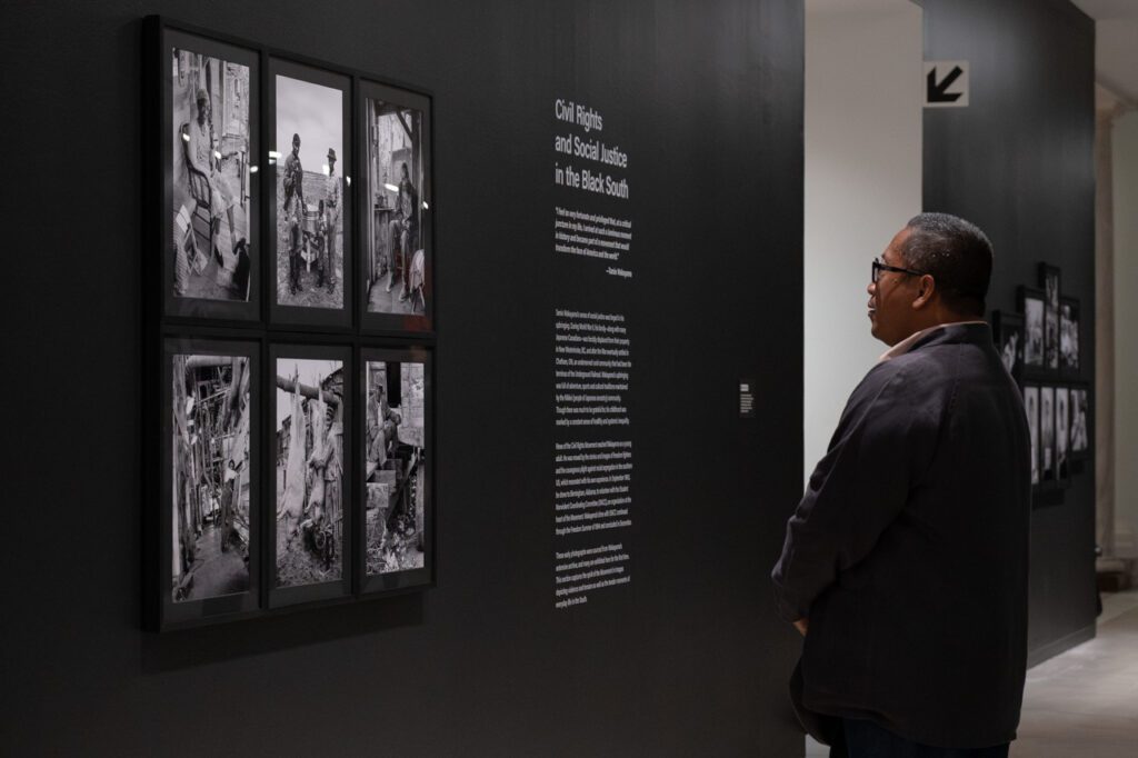 A photograph of a man standing and looking at a black wall with a series of 6 framed black-and-white photographs on it