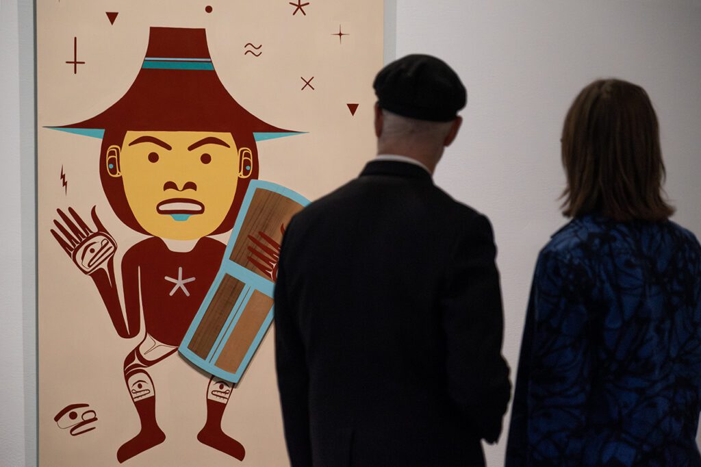 A photograph of two people, a man and a woman, from behind, looking at a painting by Bracken Hanuse Corlett of a man waving and holding a shield.