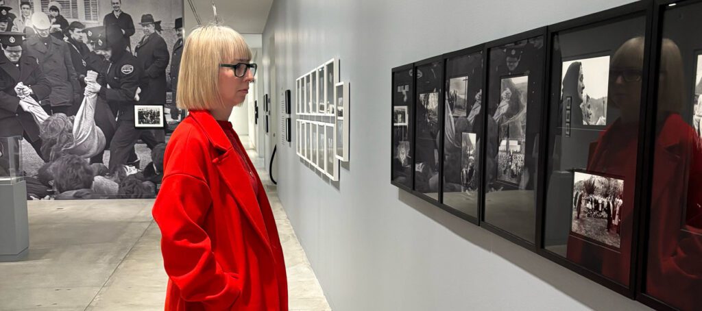 A photograph of artist Karin Bubaš from the side, wearing a bright red coat, standing in the exhibition Enemy Alien: Tamio Wakayama and looking at black and white photographs on the wall.