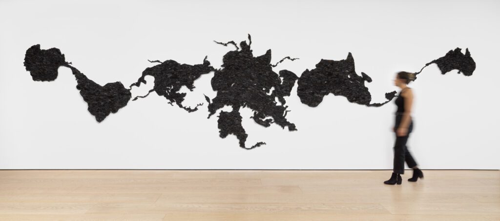 Installation view of Teresita Fernández's work Island Universe 2, made entirely of charcoal. The installation looks like a distorted map of the world, spread across one white wall. A blurred person walks in front.
