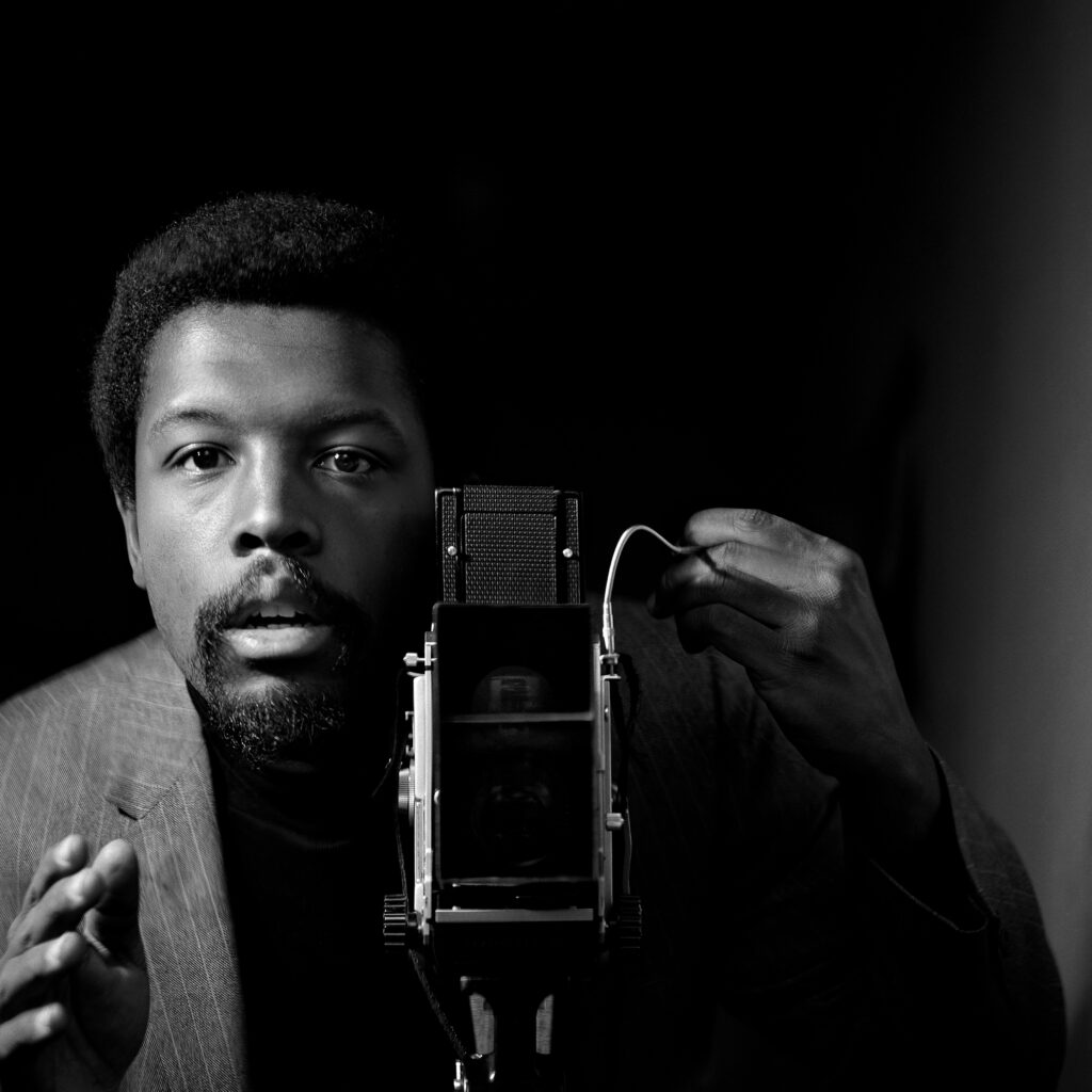 A black-and-white photograph of a Black man from the torso up standing behind a camera and looking out at the viewer. His hand is raise in the left side of the image, as if directing the scene.