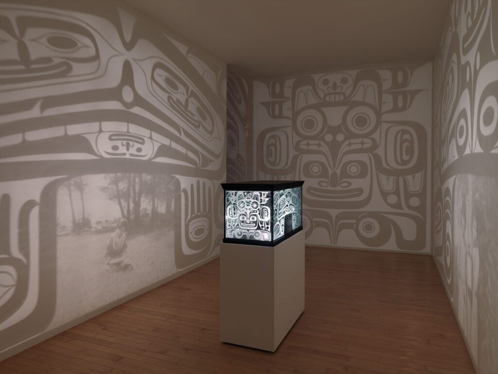 Installation by Marianne Nicolson with an object on a plinth in the centre of a room. The object looks like a bentwood box, and it shines light out on to the walls around it.