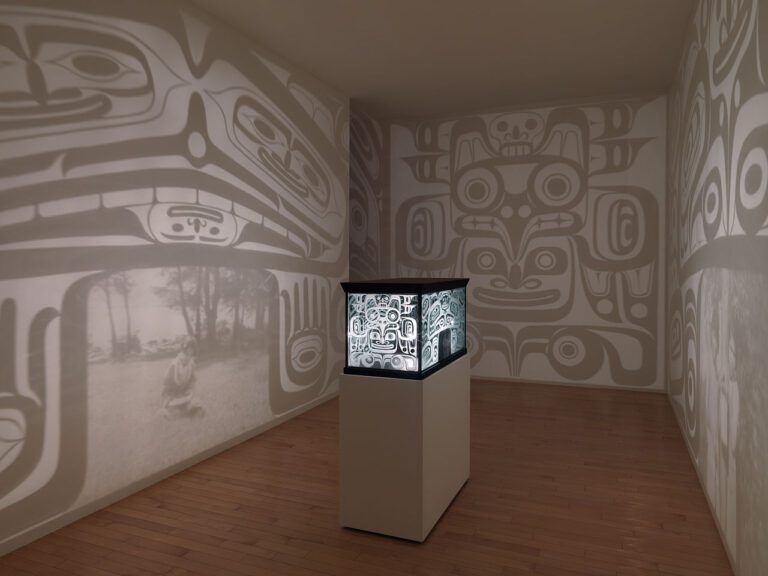 Installation by Marianne Nicolson with an object on a plinth in the centre of a room. The object looks like a bentwood box, and it shines light out on to the walls around it.