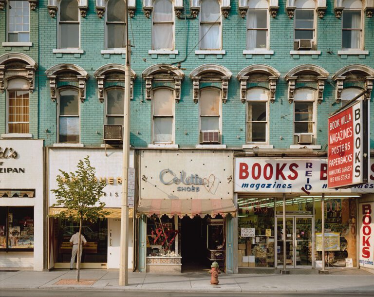 A colour photograph by Stephen Shore of a turquoise building with many windows and shop fronts, including a bookstore.