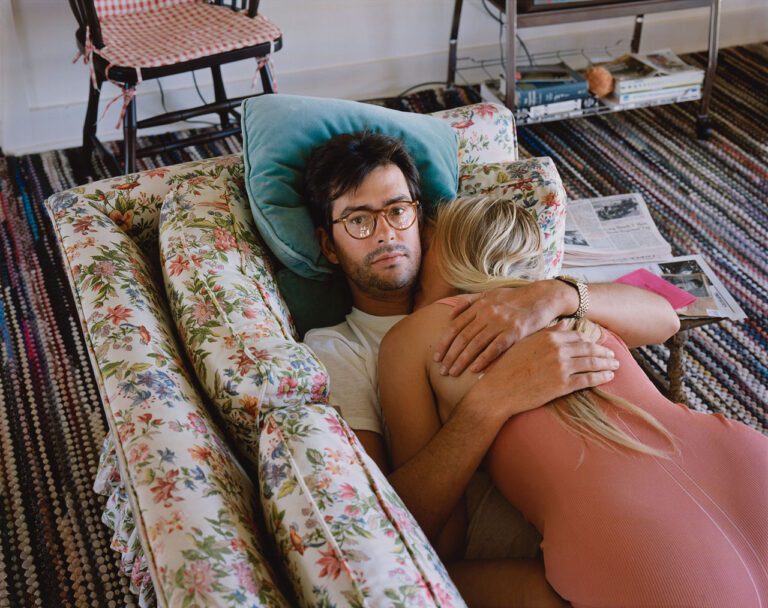 A colour photograph by Stephen Shore of a man with glasses lying on a couch looking directly at the camera. A woman with blond hair wearing a pink onesie is lying facedown on him.