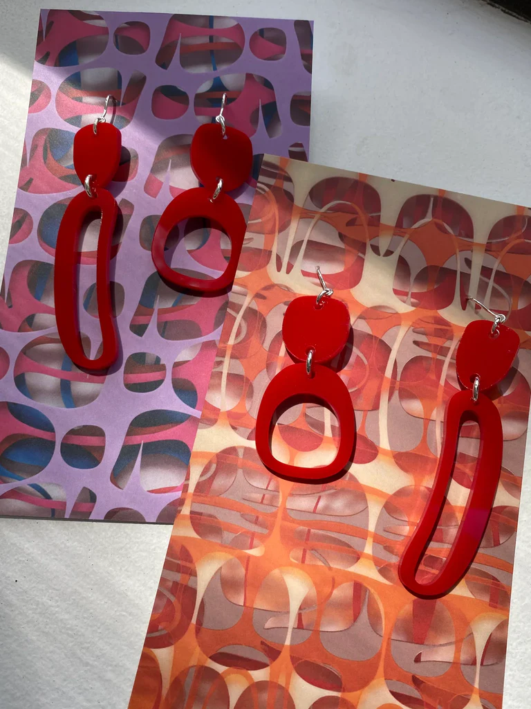 A product photograph of mini mixed ovoid earrings in red. One is on a purple colourful background and the other is on an orange colourful background.