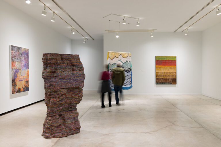 Installation view of 4 artworks by Emily Hermant in the exhibition Vancouver Now!. At the centre of the image, a couple, a man and a woman, look at the back of an hanging, tapestry-like work.
