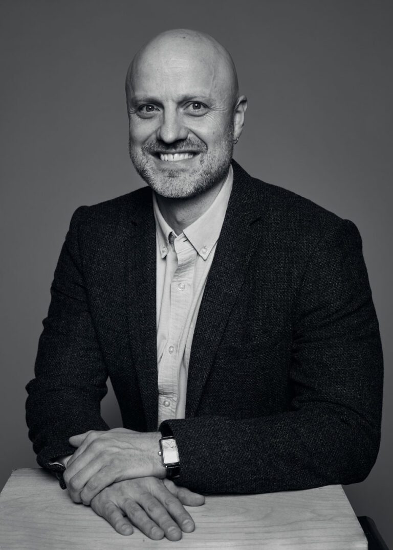 A black-and-white headshot of Grant Mosby, a white man in a black suit jacket, seen from the waist up. His hands are out in front of him.