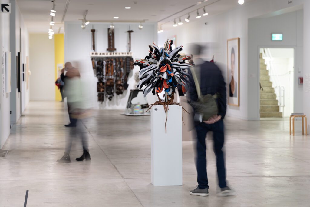 A photograph of two blurred people looking at the exhibition Highlights from the Collection. One man in the foreground looks at a sculptural work by Brian Jungen made up of sneakers, and in the back we see a large, brown structure that looks like a de-constructed piano hanging on the wall.