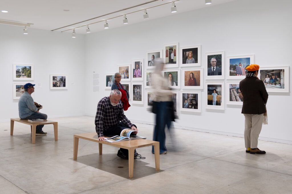 A photograph of a white-walled Gallery with white framed colour photographs on the wall. People stand and look at the photos. Others sit and look or read materials on two benches in the middle of the room.