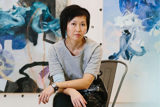 A photograph of artist Vanessa Lam sitting on a chair in her studio with large scale, abstract paintings in blue hues behind her.
