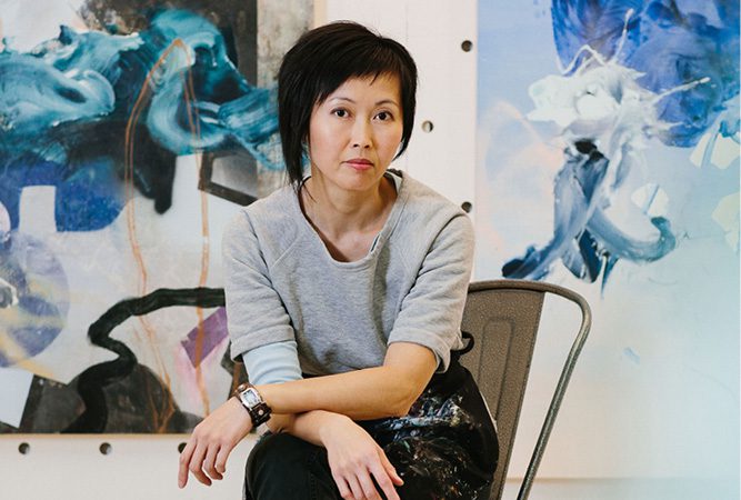 A photograph of artist Vanessa Lam sitting on a chair in her studio with large scale, abstract paintings in blue hues behind her.