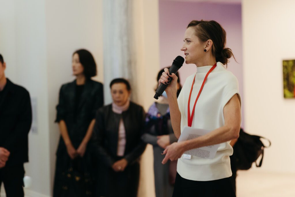 A photograph of Diana Freundl, a woman with blond hair, giving a tour. She is wearing a white shirt. Her hair is in a bun. We see people standing behind her.