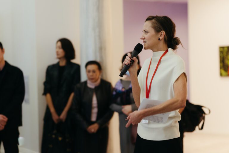 A photograph of Diana Freundl, a woman with blond hair, giving a tour. She is wearing a white shirt. Her hair is in a bun. We see people standing behind her.