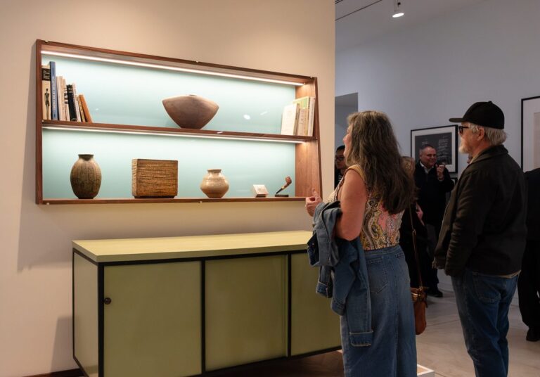 A photograph of a woman and man looking at a display in the Gallery. There is a buffet table, and above are shelves with pottery and books displayed.