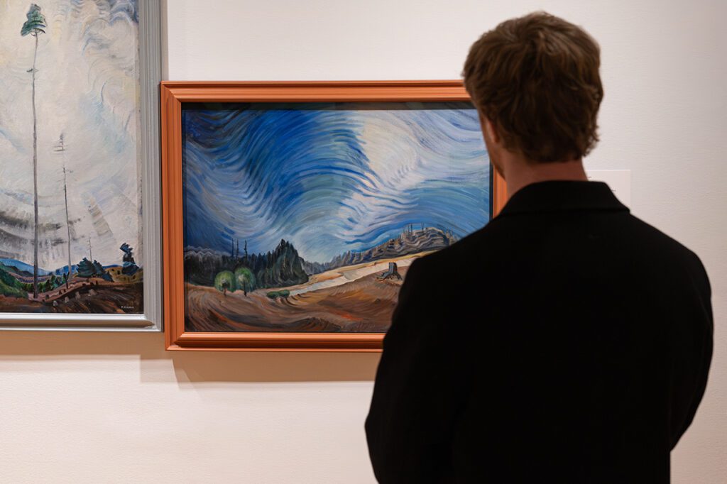 A photograph of a man from behind, from the torso up, looking at a painting by Emily Carr of a horizontal landscape.