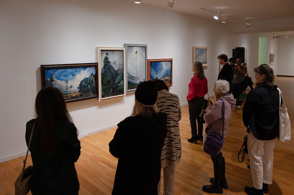 A photograph of a group of people standing in the Gallery looking at a wall of Emily Carr paintings.