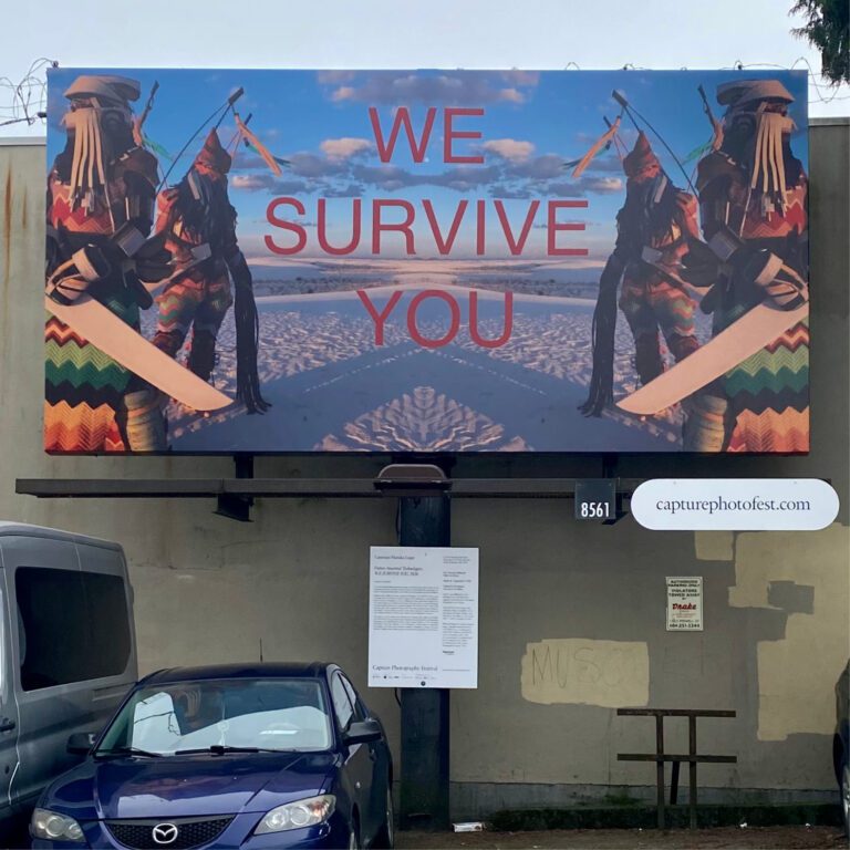 A square photograph of a billboard outside in a parking lot. The billboard features an artwork by Cannupa Hanska Luger with 4 people, 2 on either side of the billboard, dressed in futuristic looking traditional Indigenous clothing, and in the middle the text "WE SURVIVE YOU" In red all caps.
