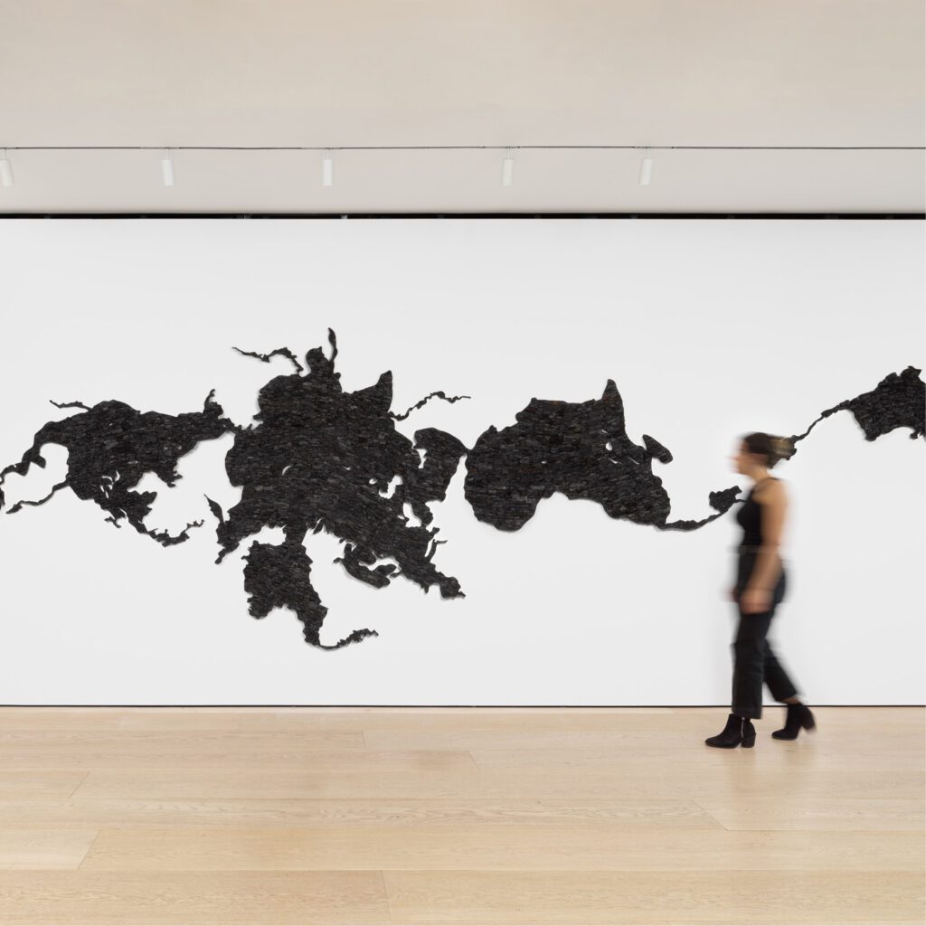 Installation view of Teresita Fernández's work Island Universe 2, made entirely of charcoal. The installation looks like a distorted map of the world, spread across one white wall. A blurred person walks in front. This is a square crop.