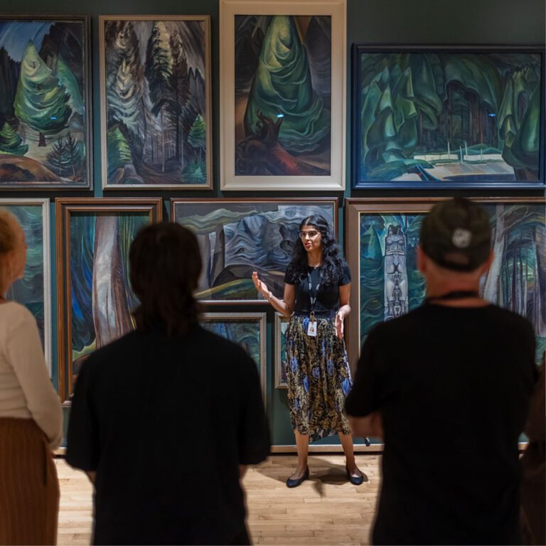 A group of men and women visit the Gallery and enjoy a guided tour. They are standing with their backs to us, looking at a dark green wall with Emily Carr paintings of the forest on it. A young woman tour guide stands in front of the paintings gesturing and looks back at the group.