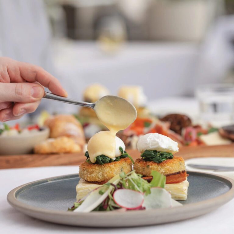 A square image of a hand pouring hollandaise sauce onto eggs benedict on a plate.