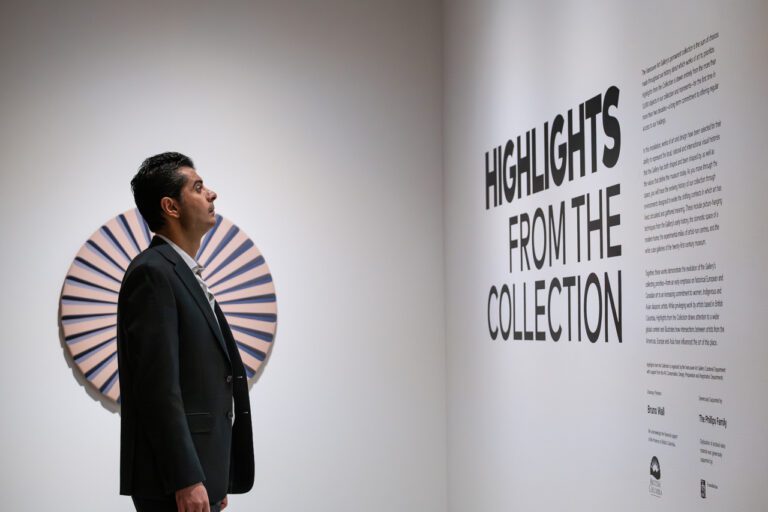 A photograph of a man from the side standing in front of the title wall in an exhibition space. The exhibition title appears on the grey wall in black: Highlights from the Collection. Behind the man is a circular sculpture on the wall in pink and purple.