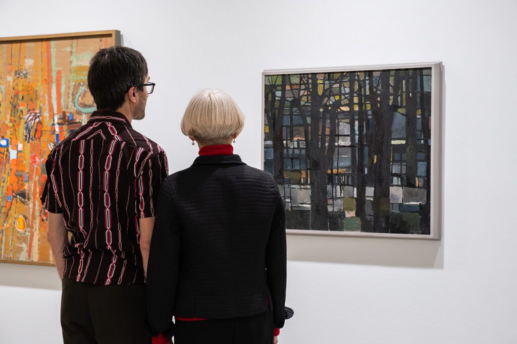 A photograph of two people, a mand and a woman, looking at paintings on a white gallery wall.
